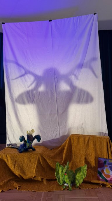 Scena teatralna, a za powieszonym prześcieradłem cień kobiety z porożem jelenia